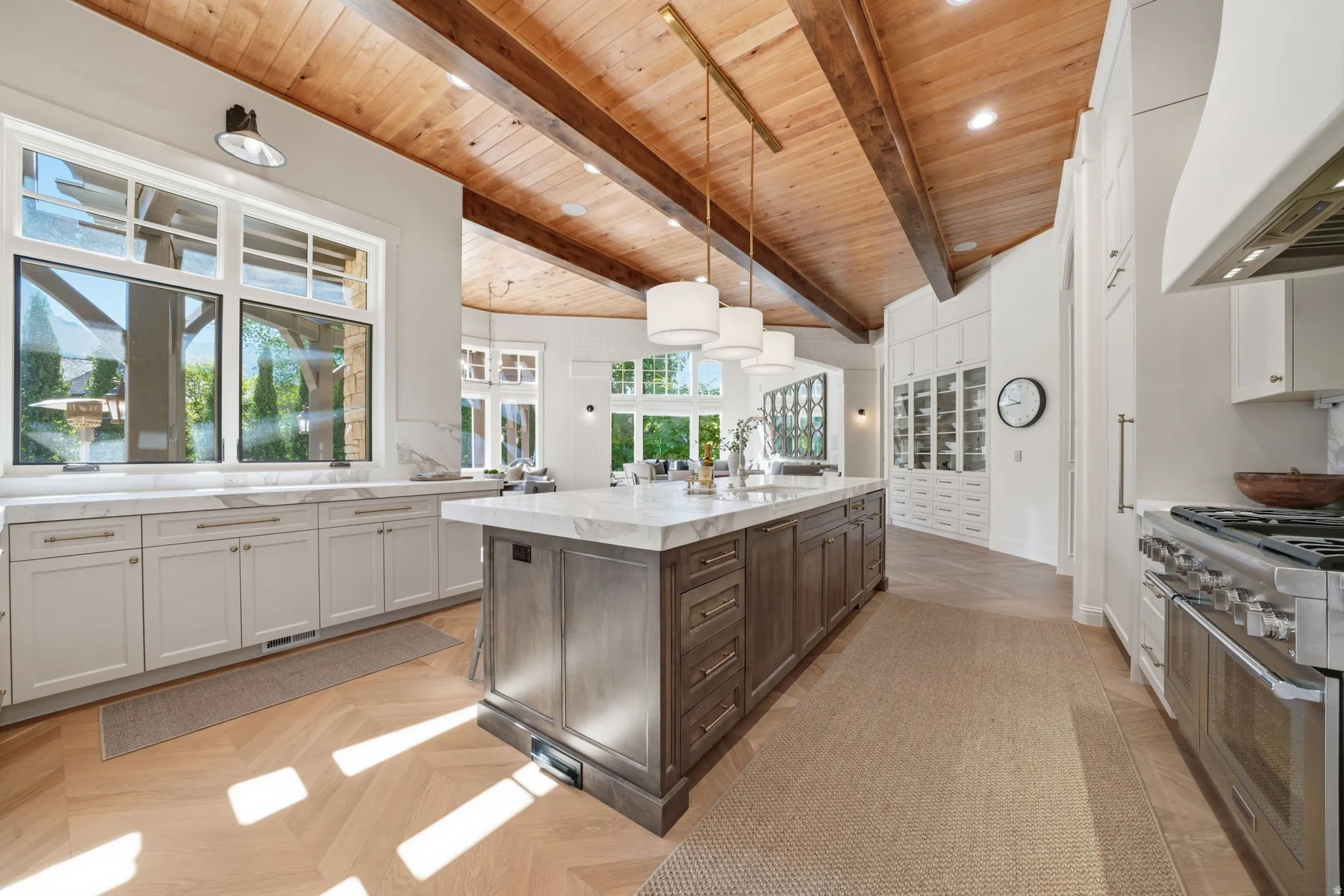 Expansive kitchen featuring a large central island with a light-toned countertop, wood-finish flooring in a herringbone pattern, exposed wood beam ceiling, white cabinetry with silver hardware, and a stainless steel range