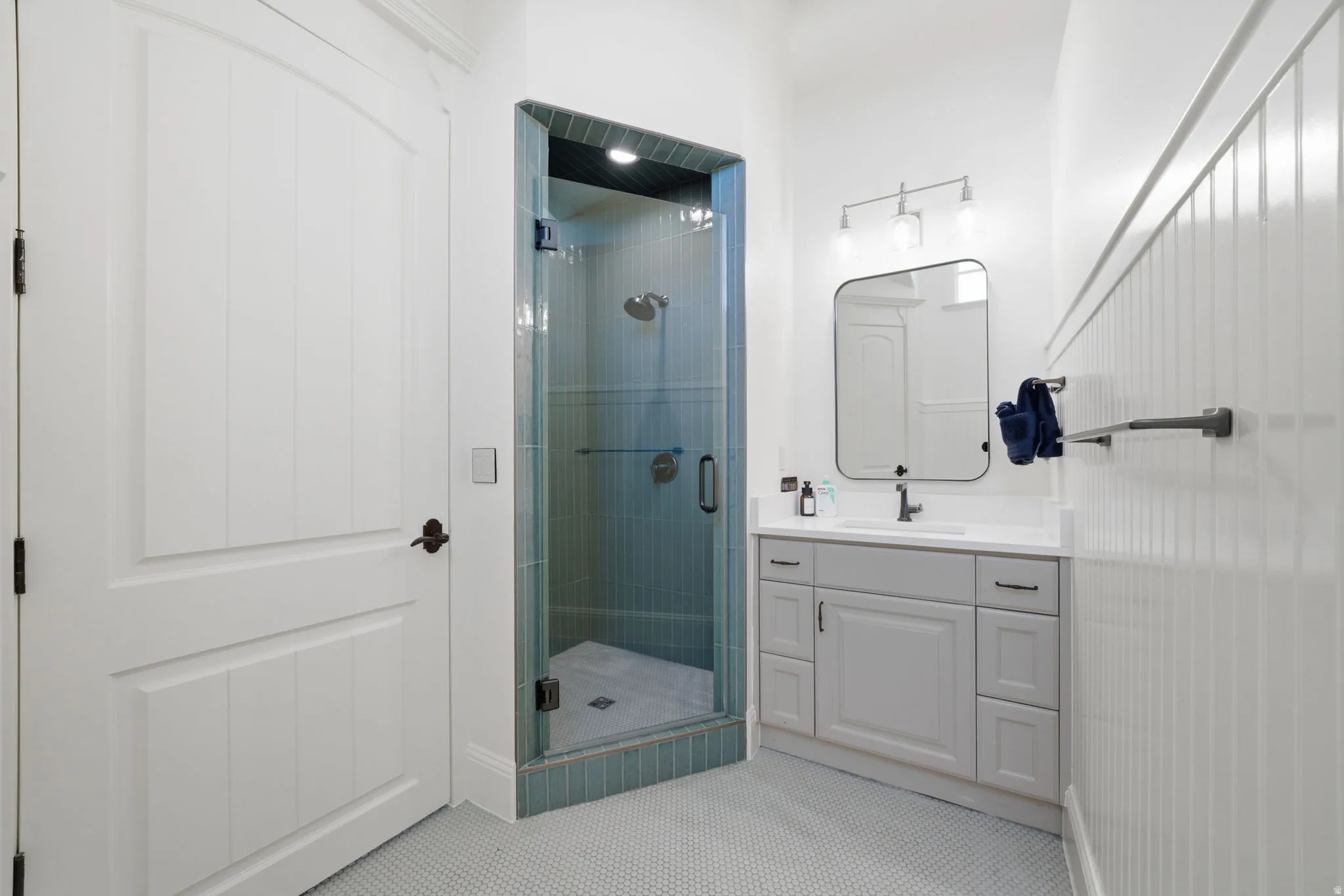 Full bathroom featuring a glass-enclosed shower with turquoise tile, a single-sink vanity with a light gray cabinet and white countertop, and white beadboard paneling