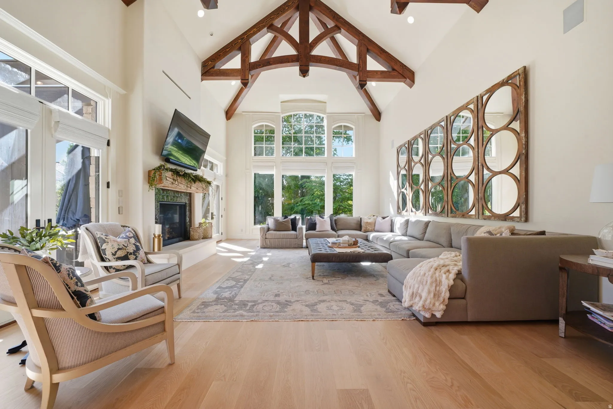 Vaulted ceiling with exposed wood beams, expansive window wall with arched transoms, light wood-finish flooring, stone fireplace with a tile surround, and multiple sets of French doors