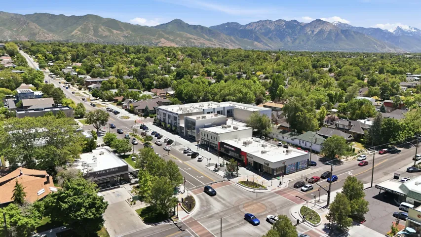 Aerial perspective of a commercial intersection with multiple businesses and residential properties, framed by distant mountains and abundant green trees