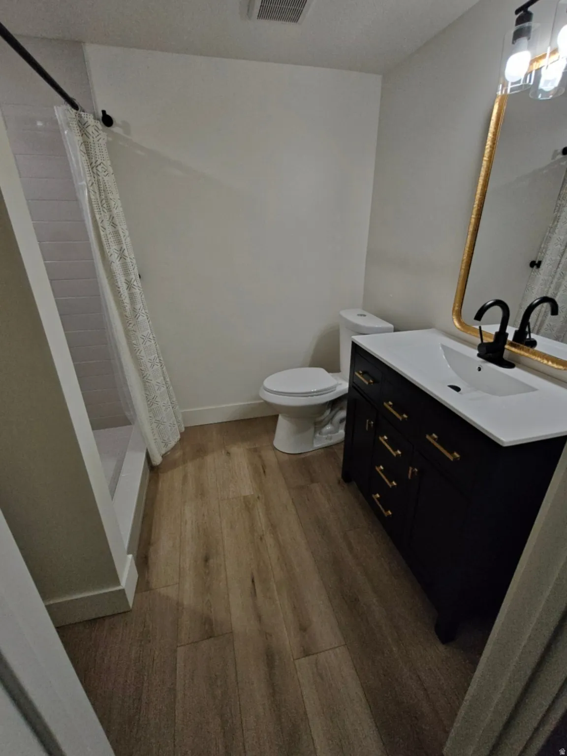 Bathroom featuring wood-finish flooring, a dark vanity with gold-tone hardware, a white toilet, and a shower with white subway tile