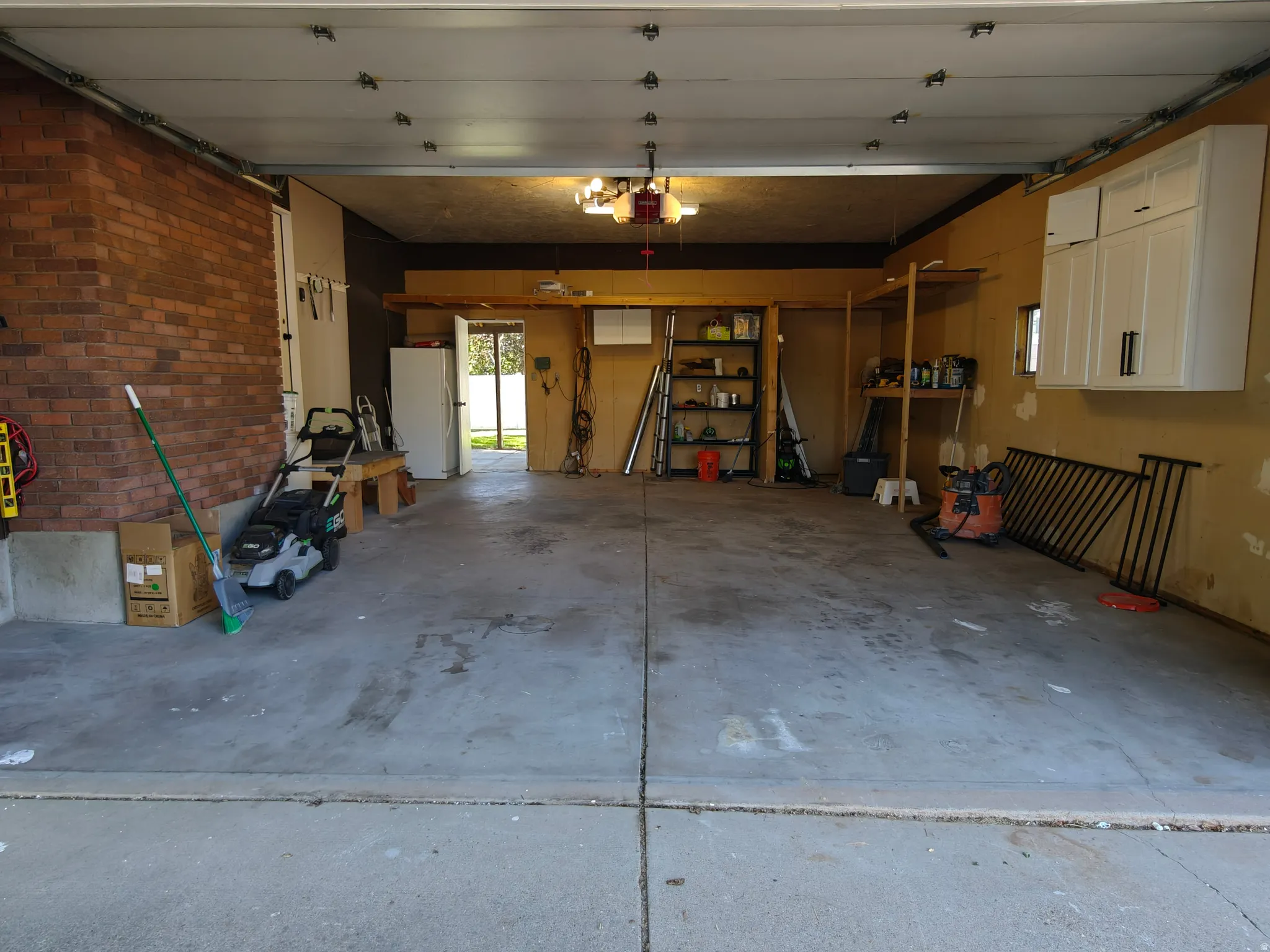 Spacious garage with exposed brick wall, concrete flooring, overhead lighting, built-in shelving, and white wall-mounted cabinetry