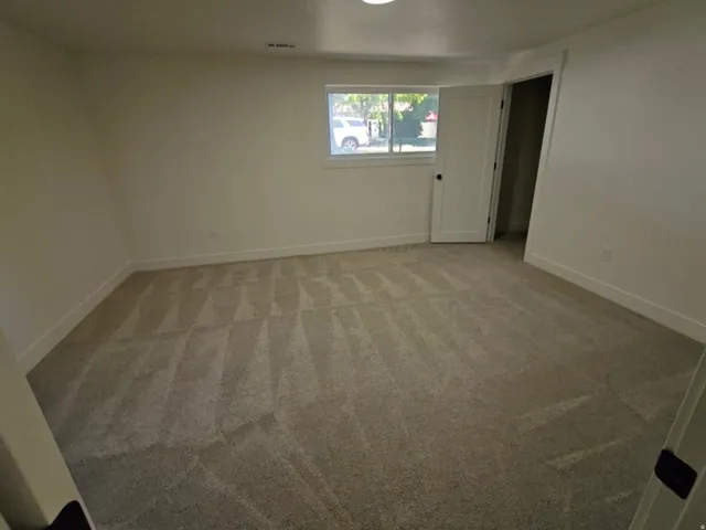 Spacious room featuring new carpeting, white baseboards, and a window with natural light
