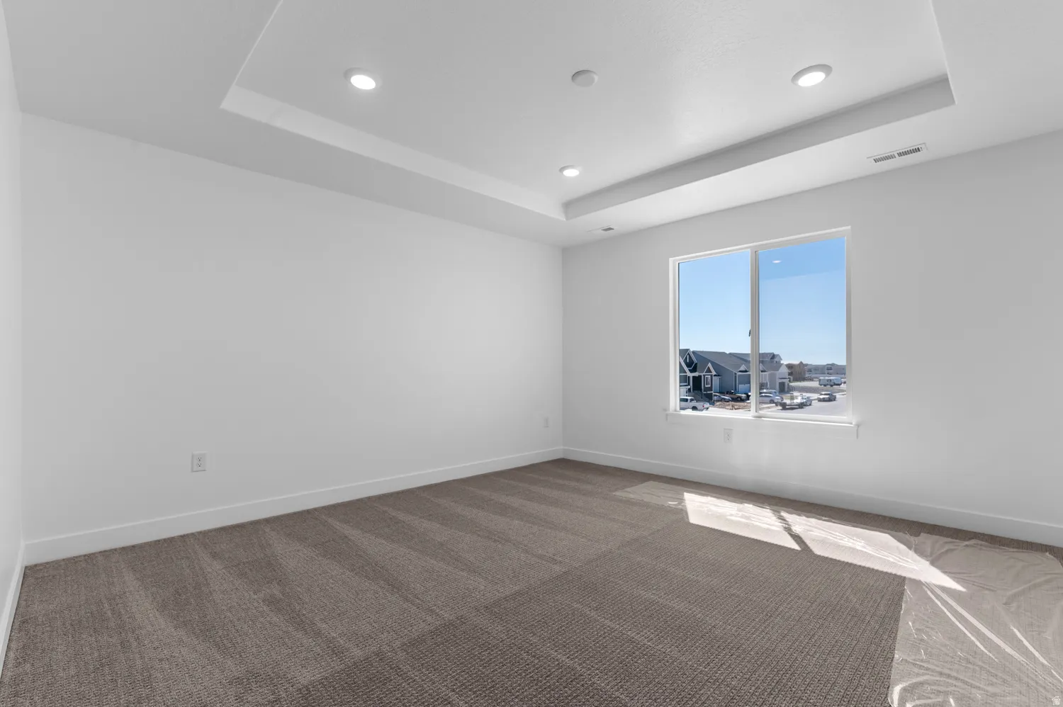 Recessed lighting illuminating a tray ceiling, expansive window, plush carpet, and white baseboards
