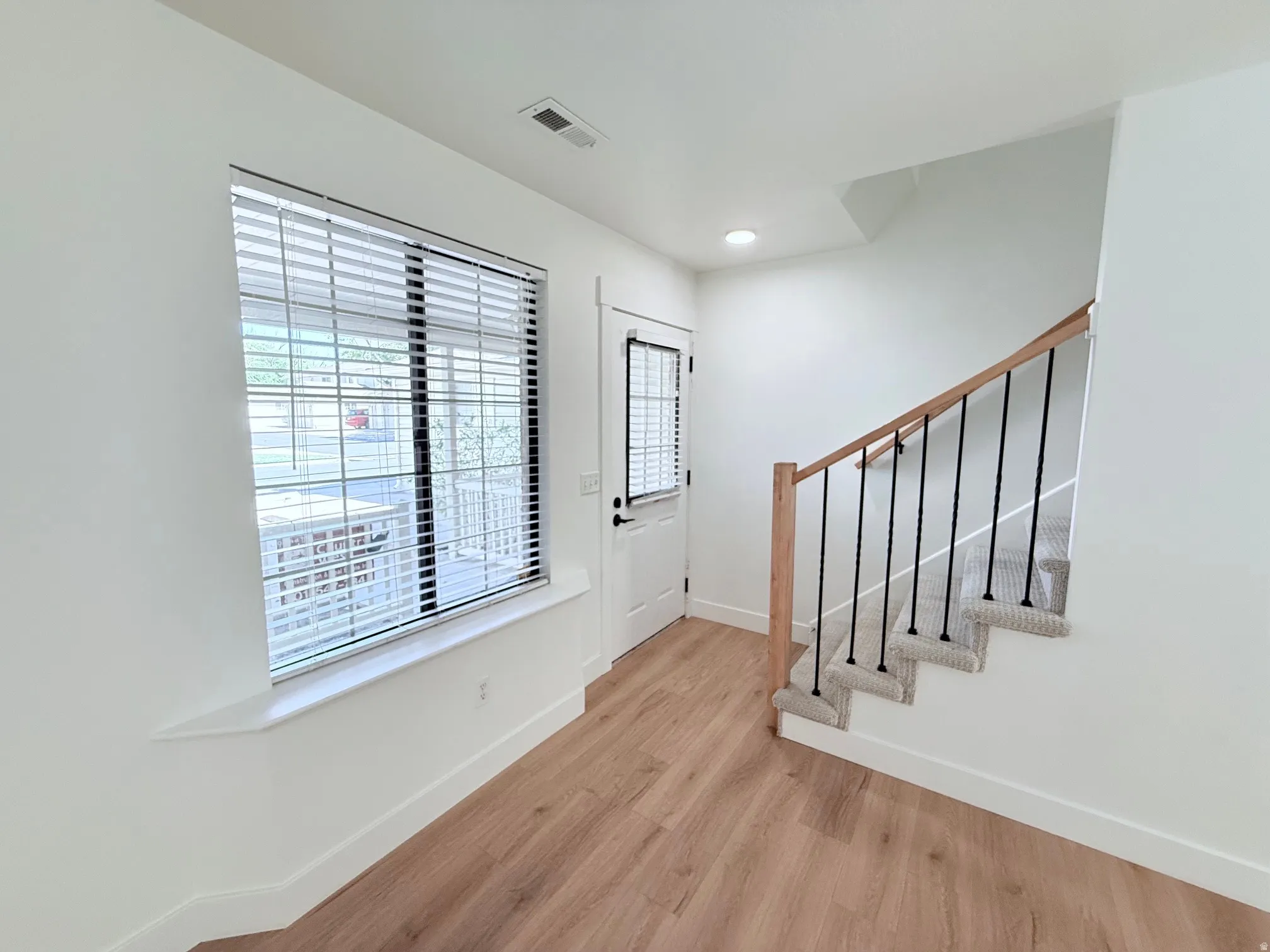 Wood-finish flooring, white walls, staircase with carpeted treads, wood handrail, black balusters, and a window with blinds