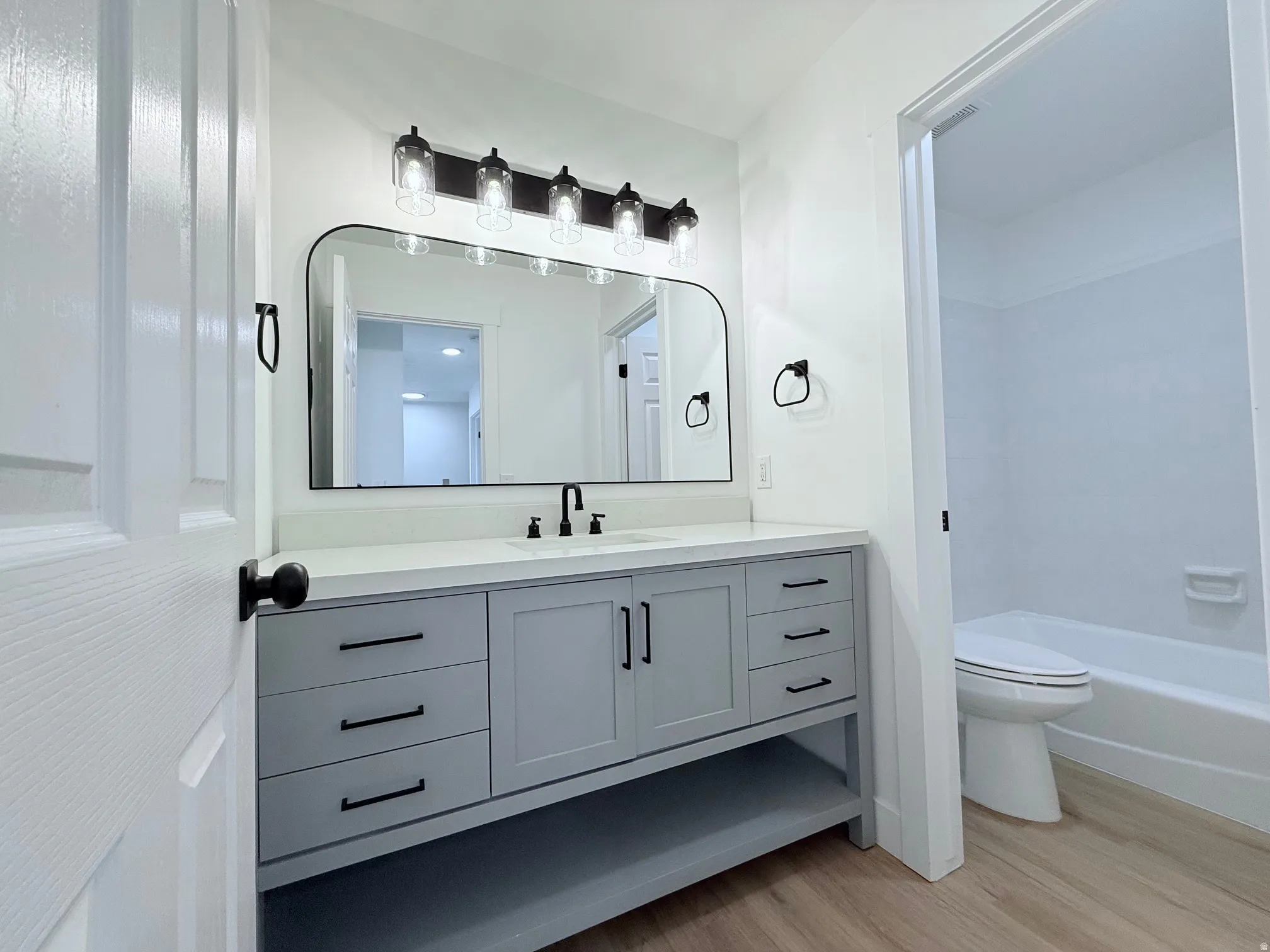 Light wood-finish flooring, a white vanity countertop with an integrated sink, a large arched mirror, and a contemporary five-light fixture with clear glass shades