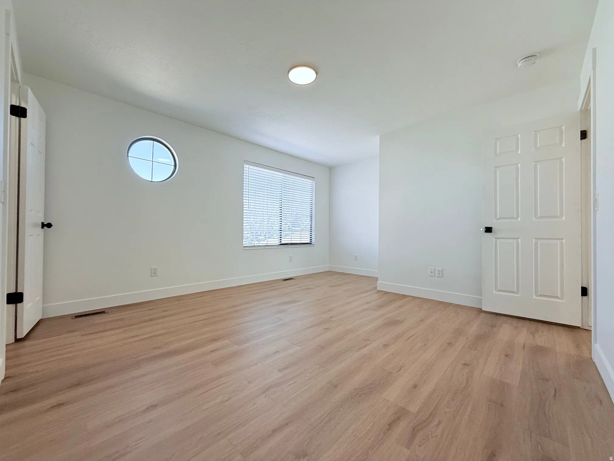 Wood-finish flooring, white walls, circular accent window, rectangular window with blinds, and a flush mount ceiling light
