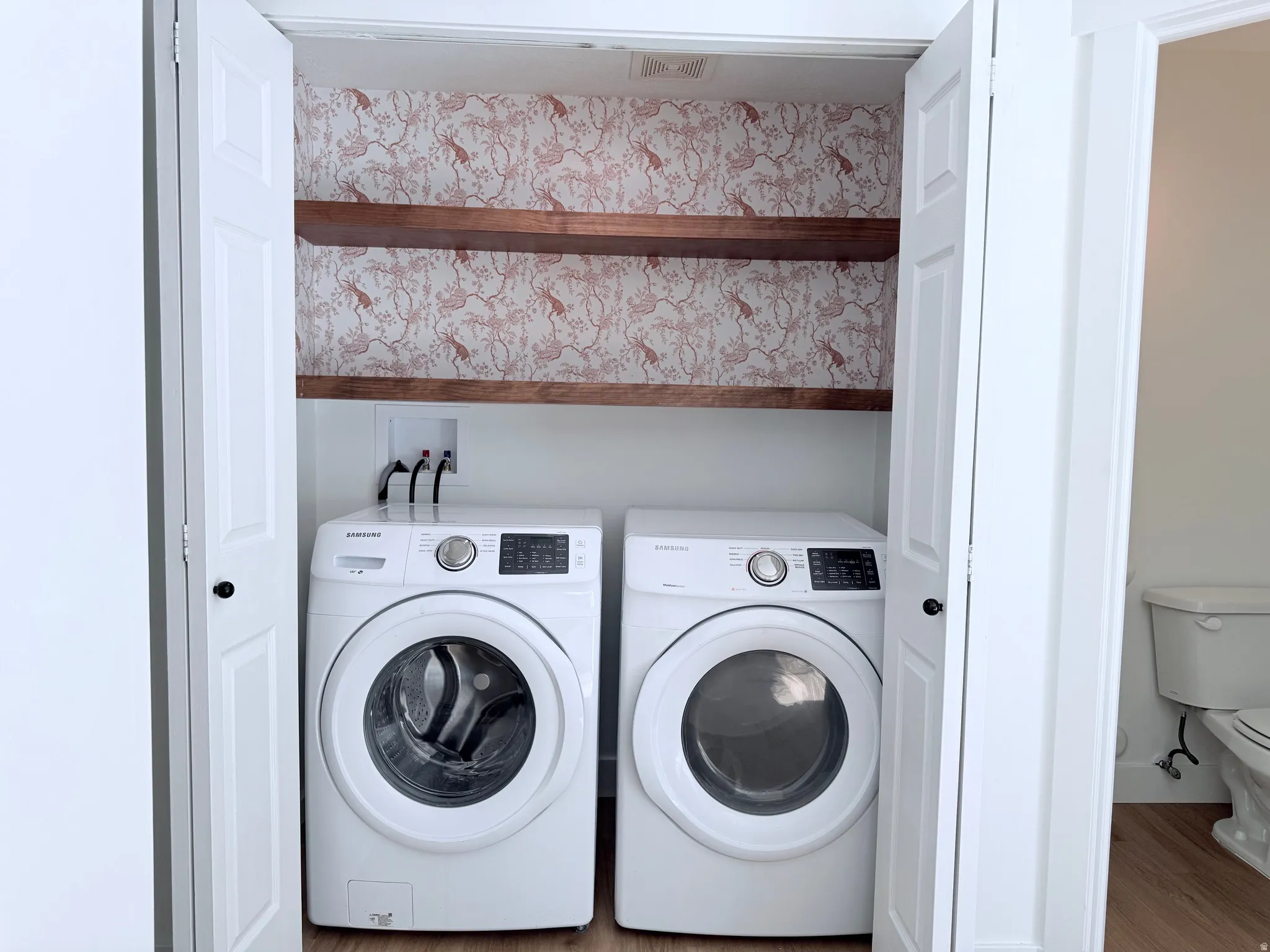 Dedicated laundry closet featuring side-by-side front-load washer and dryer with digital controls, wooden shelving above for storage, and decorative wallpapered backing