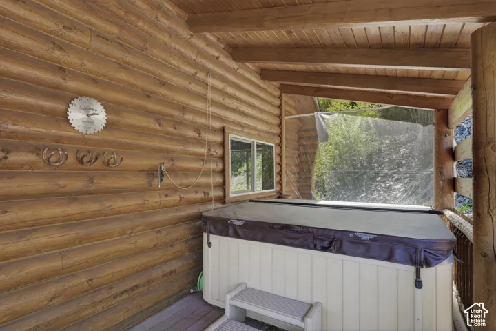 Covered exterior patio featuring a spa with cover, log walls, wood ceiling beams, and a window