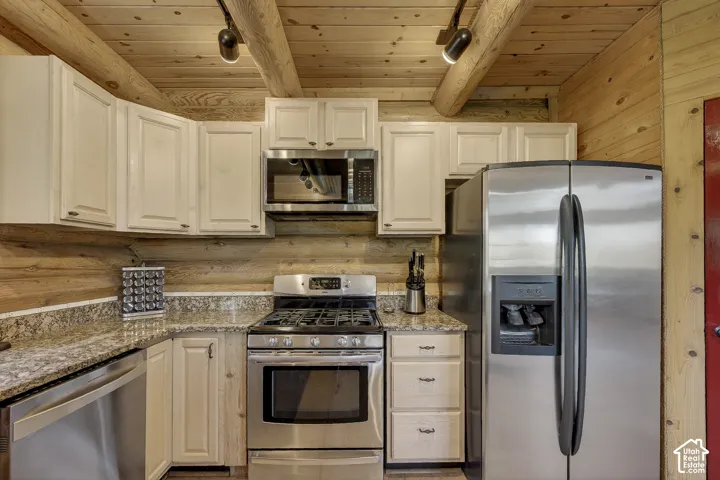 Kitchen featuring stainless steel appliances, light-toned cabinetry, natural wood-paneled walls, a wood-beam ceiling, and stone-finish countertops