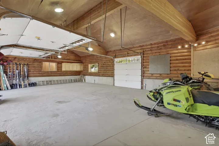 Spacious garage featuring log wall construction, exposed wood beam ceiling, concrete flooring, dual overhead garage doors, and built-in cabinetry