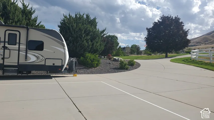 Spacious concrete driveway featuring ample parking, a separate paved pathway, a white three-rail fence, and mature trees