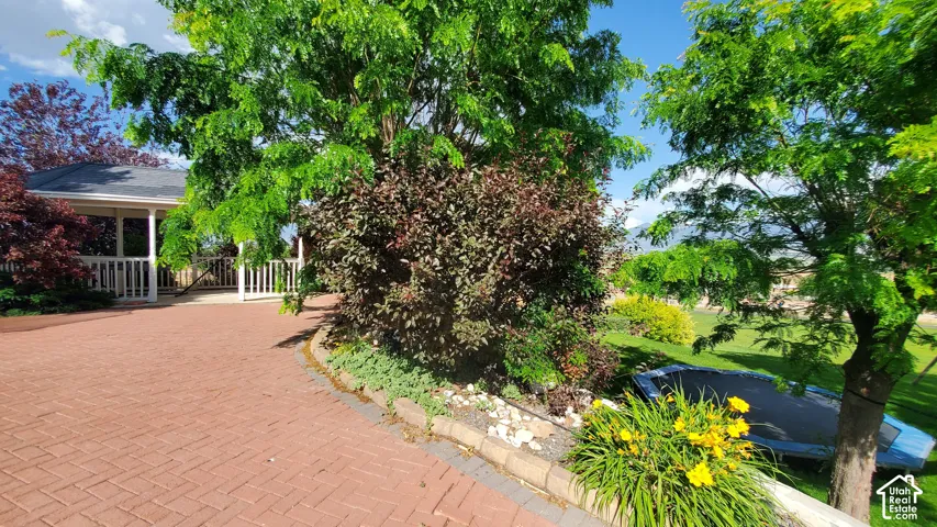 Brick paver driveway with a white-railed porch featuring a gray shingled roof, bordered by mature trees and shrubs