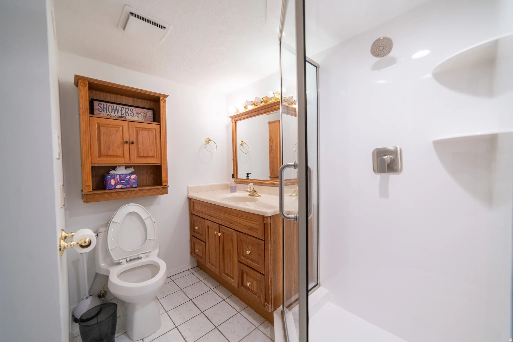Bathroom featuring a glass-enclosed shower with integrated shelving, a single vanity with a light-colored countertop, wood-finish cabinetry, a framed mirror, and white square tile flooring