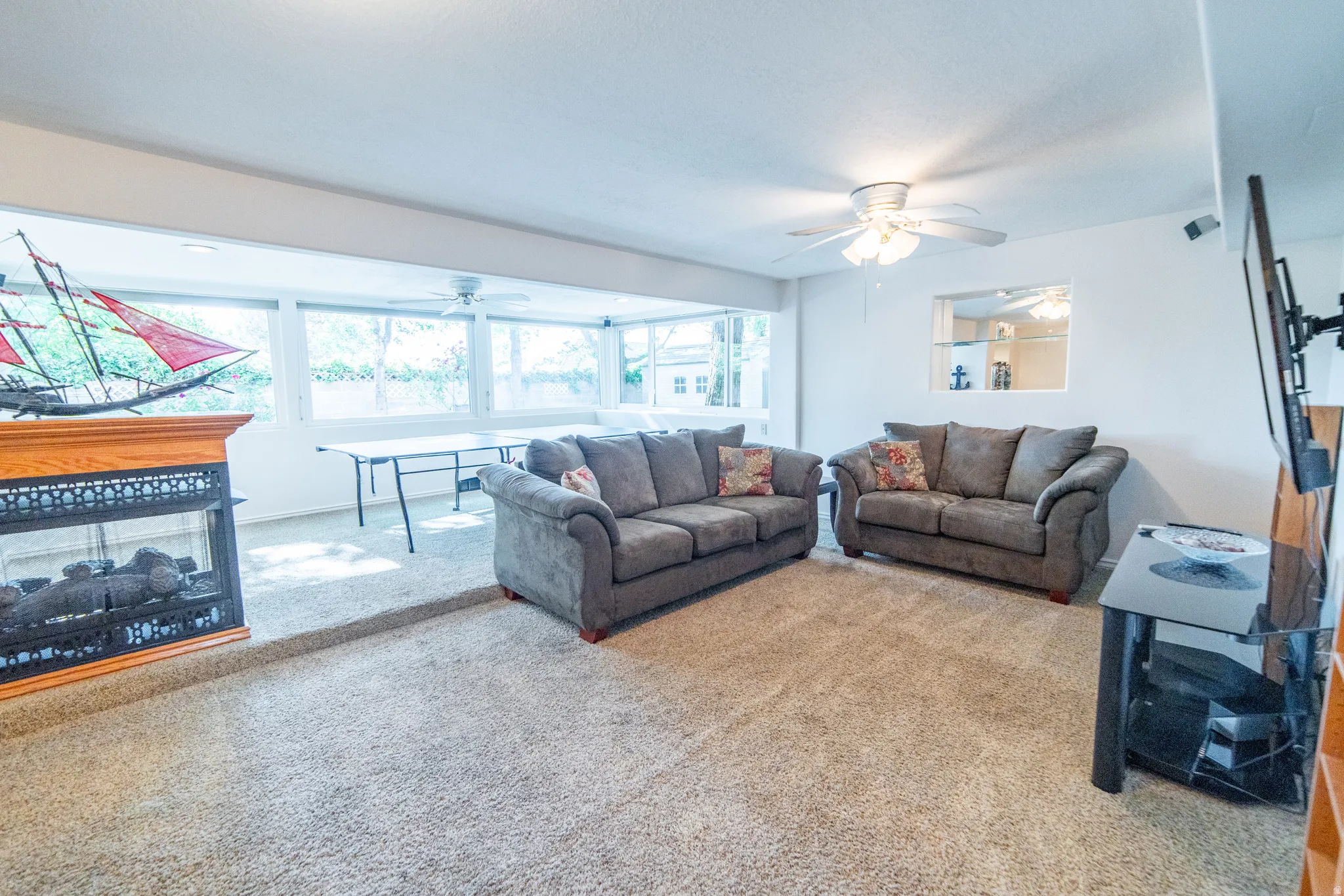 Carpeted living area featuring a fireplace with an oak mantel, a ceiling fan, and large windows providing ample natural light