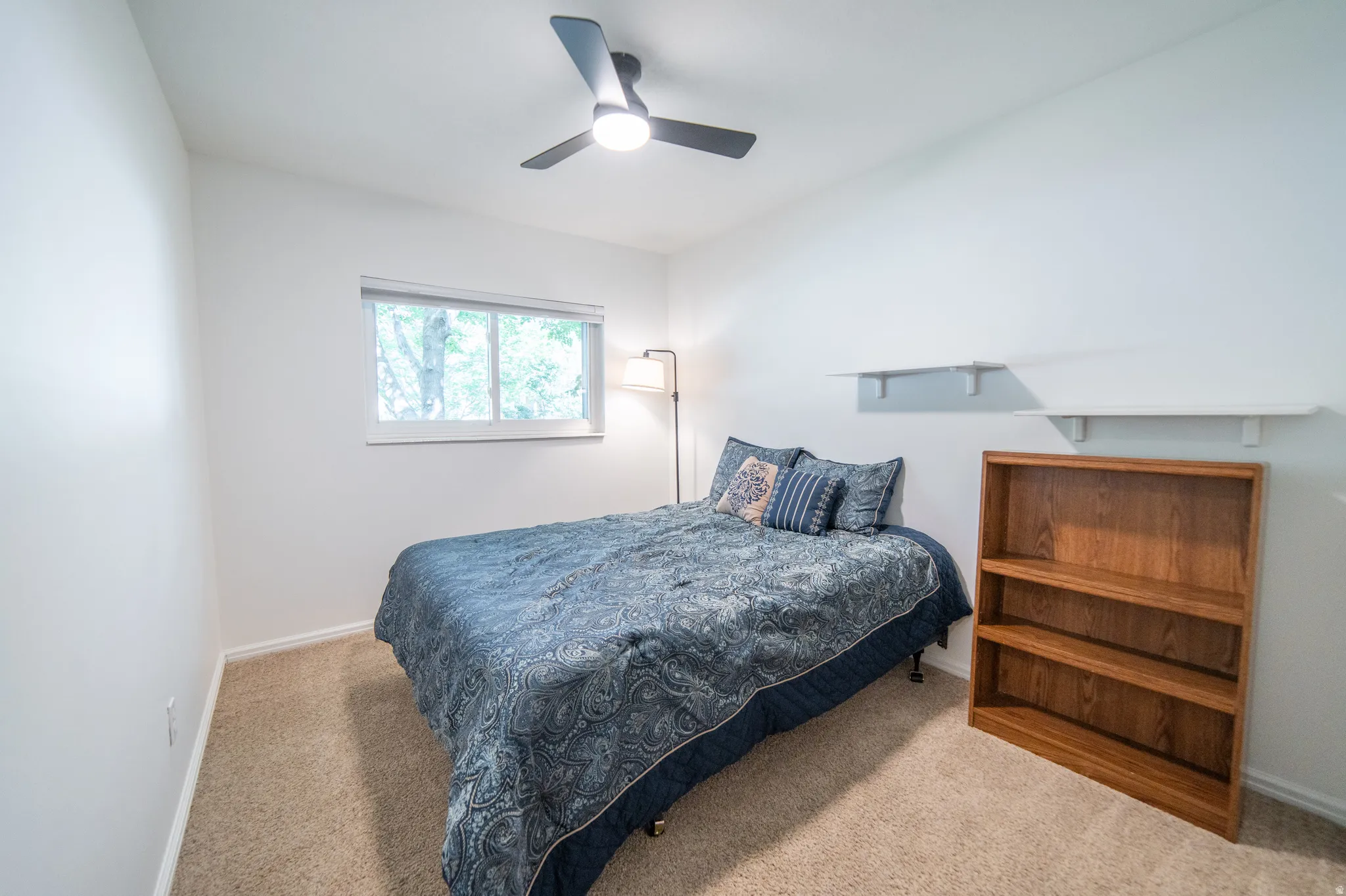 Bedroom featuring a ceiling fan with integrated lighting, a window with exterior tree scenery, light-toned carpeting, and white wall shelving
