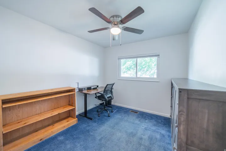 Bright interior room featuring a window with exterior tree views, a ceiling fan with integrated lighting, and blue carpeting