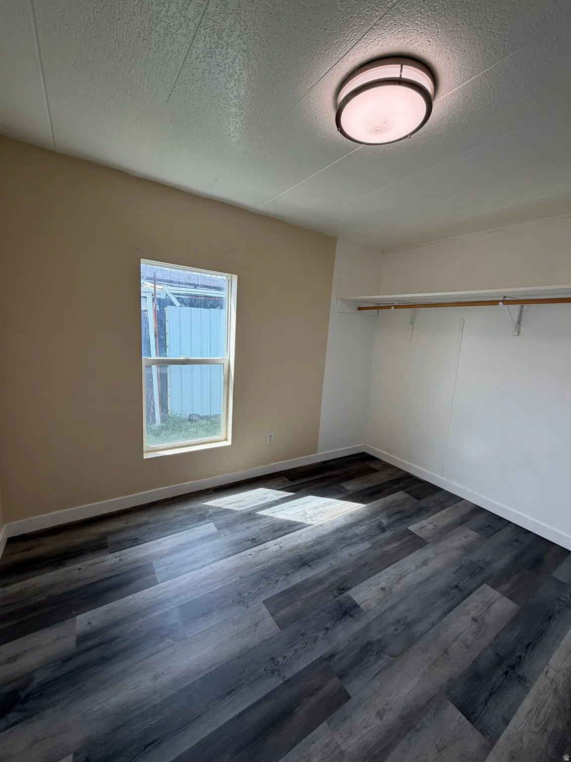Spacious room featuring wood-finish flooring, a flush-mount ceiling light fixture, a window with exterior visibility, and a built-in closet with shelving and hanging rod