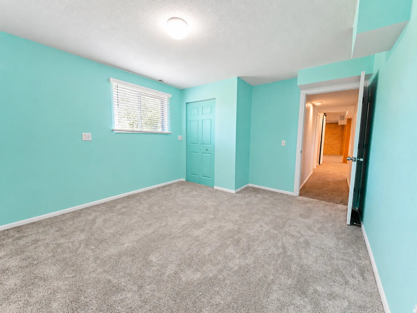 Spacious room featuring light blue walls, plush gray carpeting, a window with horizontal blinds, and a bi-fold closet door
