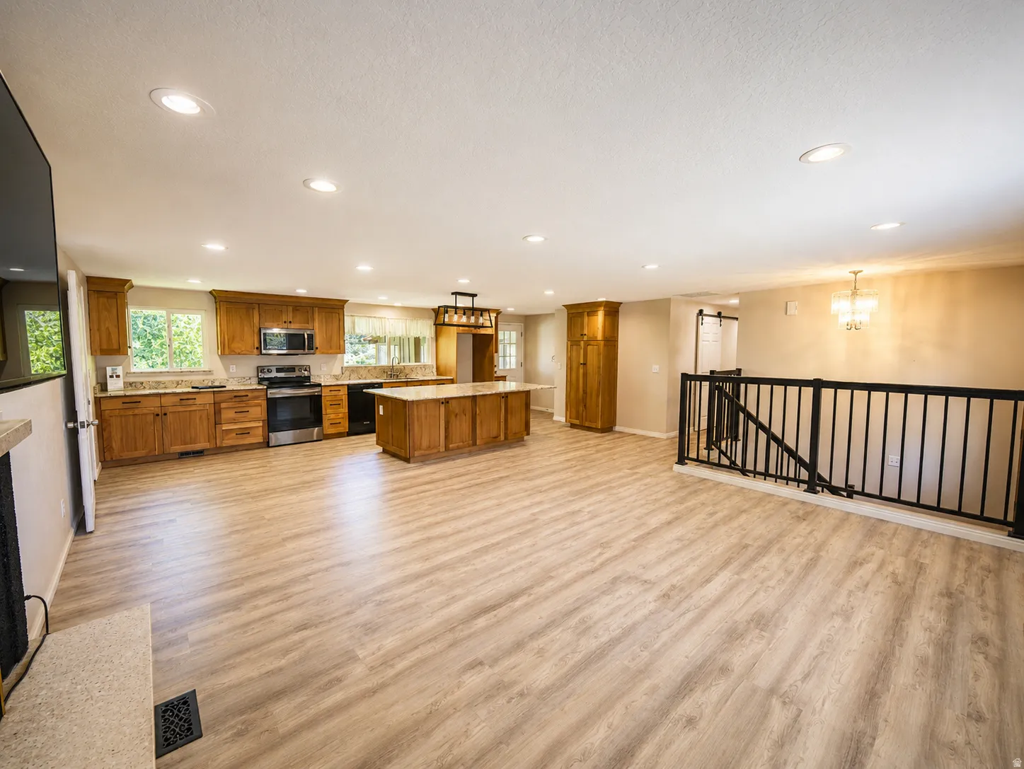Open-concept living space featuring wood-finish flooring, recessed lighting, and a kitchen with wood cabinetry, stainless steel appliances, and a central island with a stone-look countertop