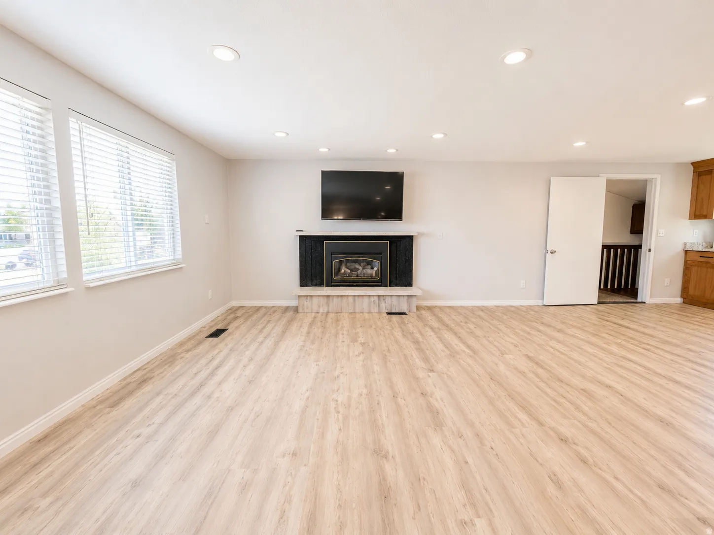 Spacious room featuring wood-finish flooring, recessed ceiling lighting, a fireplace with a black surround and stone hearth, and multiple windows with blinds