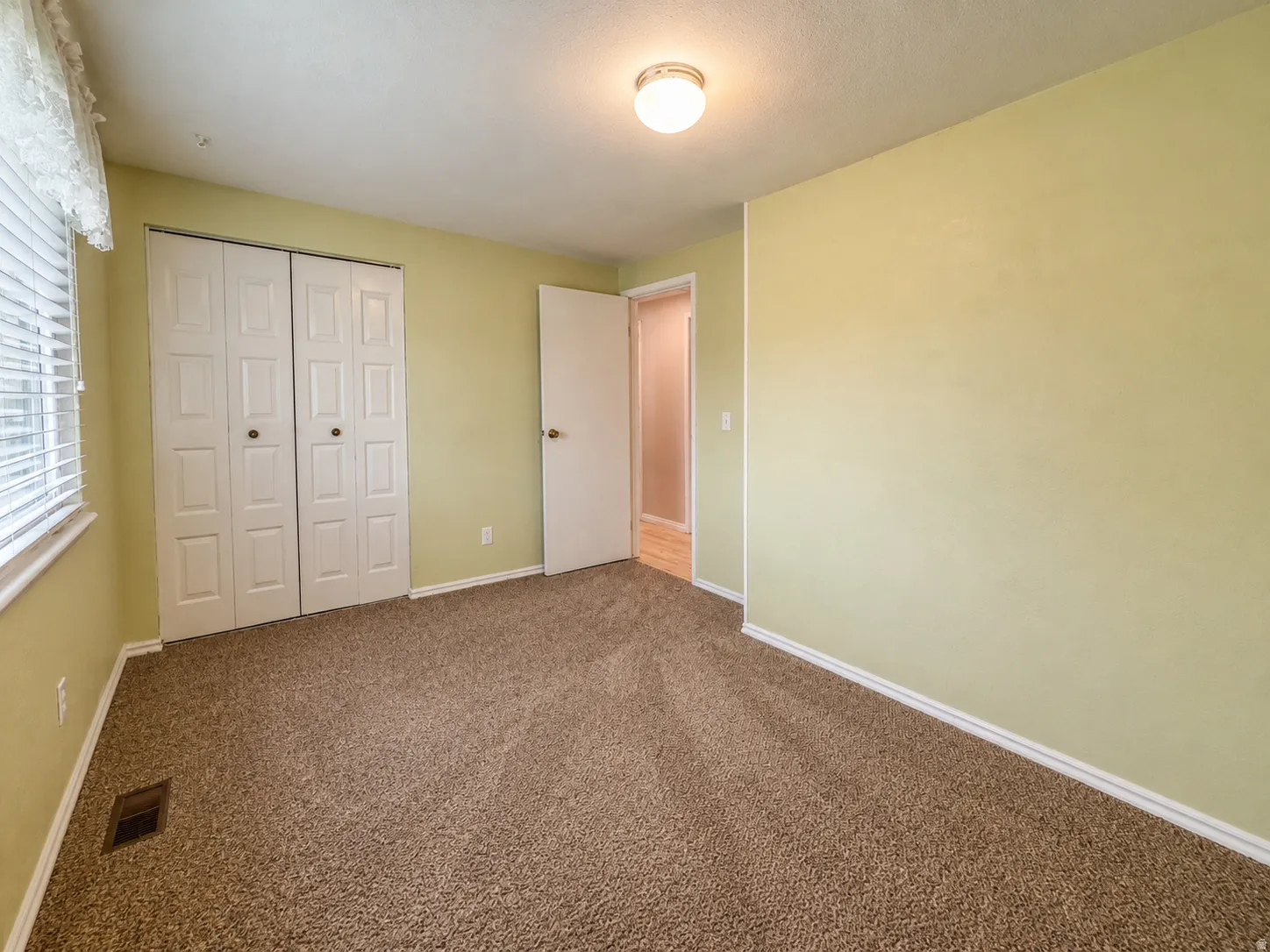 Carpeted room featuring a white paneled bi-fold closet, a single window with blinds, a white door frame, and a ceiling-mounted light fixture