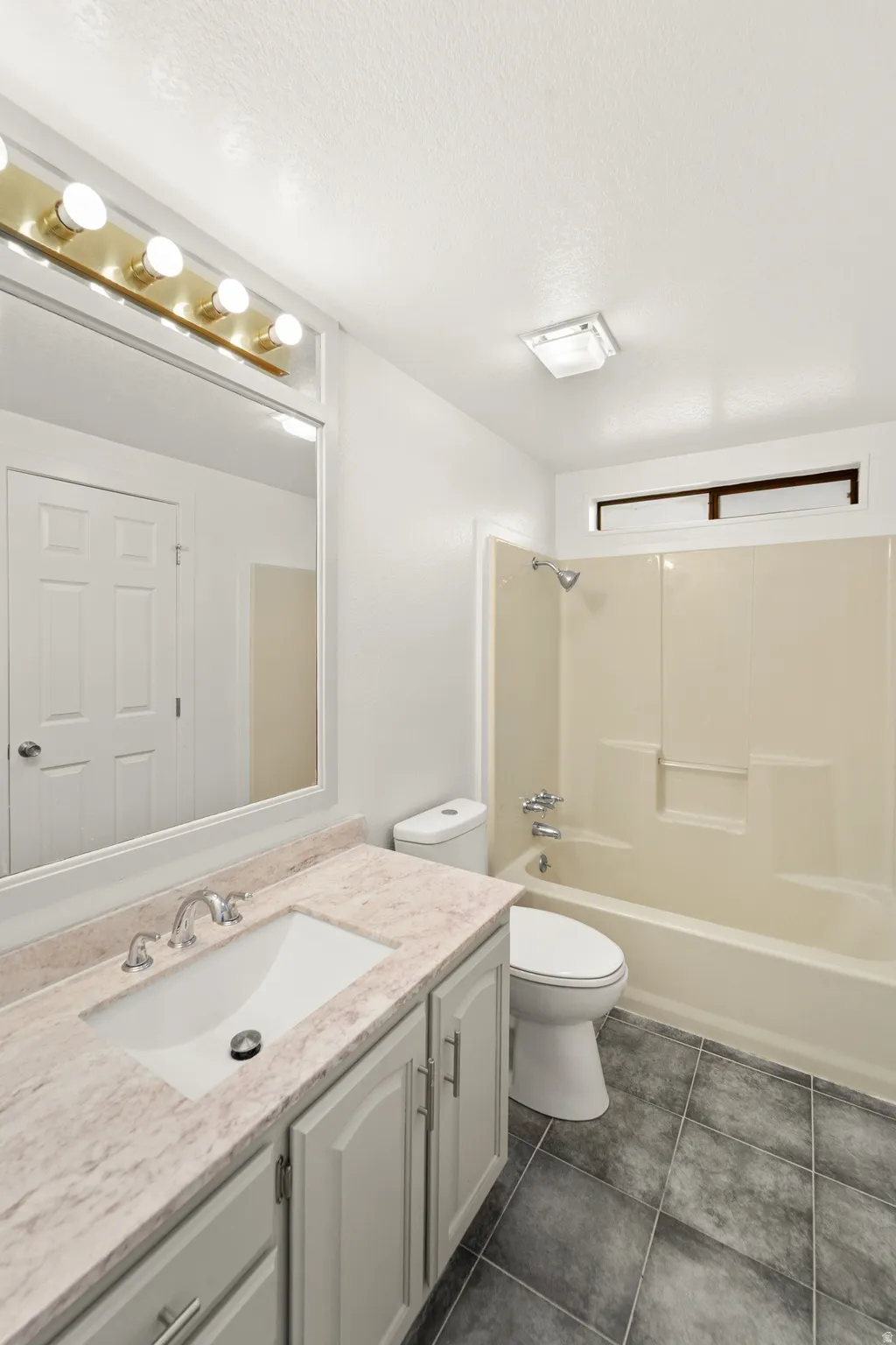 Bathroom featuring a built-in vanity with a rectangular sink and stone-finish countertop, a full-size mirror with a five-bulb vanity light fixture, a toilet, a shower-over-tub combination with a built-in niche, and dark square floor tiles