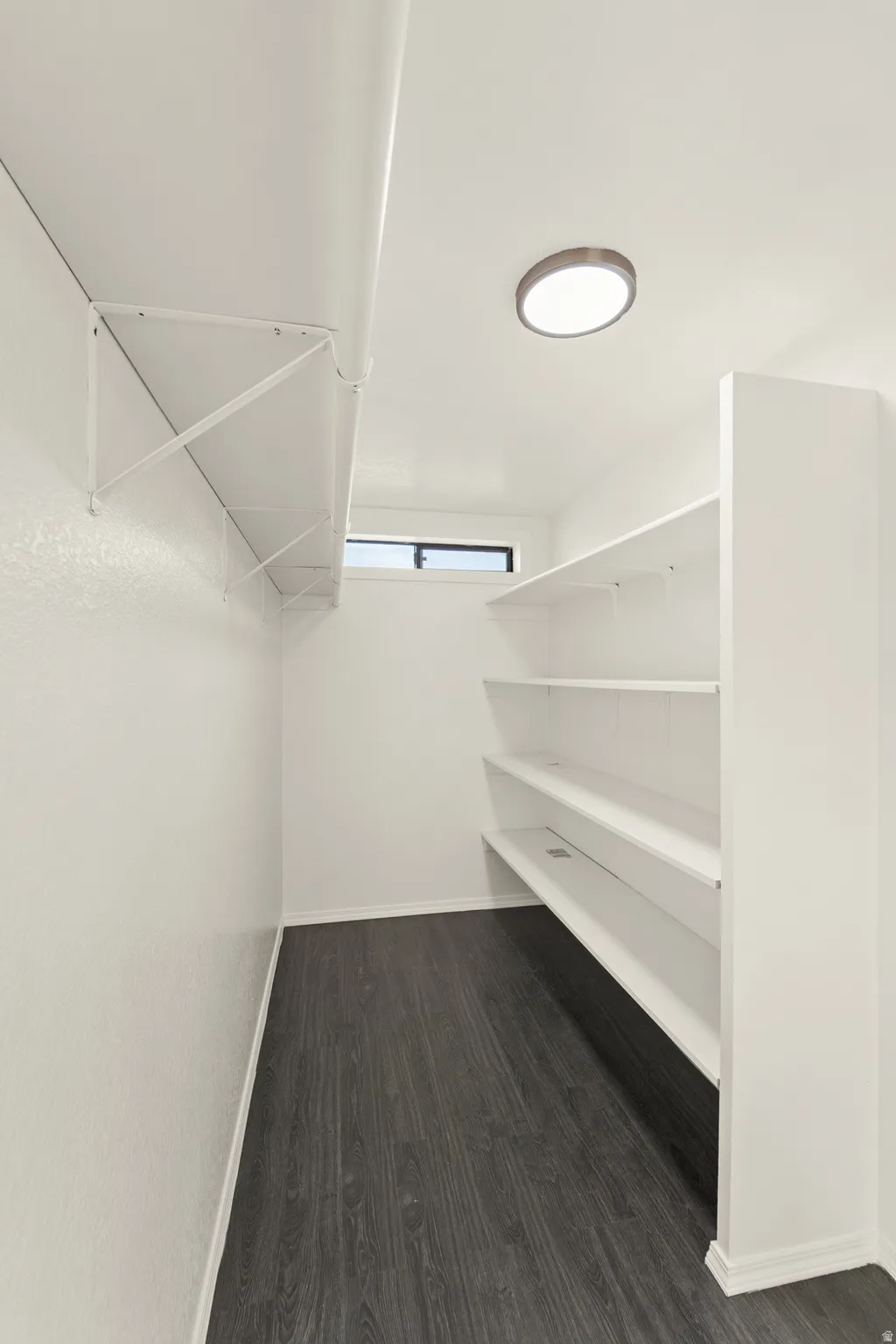 Walk-in storage area featuring multiple white shelving units, wood-finish flooring, a recessed ceiling light, and a horizontal window