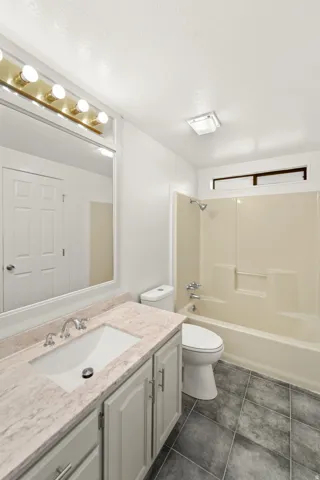 Bathroom featuring a built-in vanity with a rectangular sink and stone-finish countertop, a full-size mirror with a five-bulb vanity light fixture, a toilet, a shower-over-tub combination with a built-in niche, and dark square floor tiles