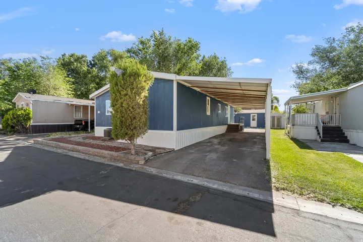 Exterior featuring blue siding with white trim, a covered carport, a stone-edged planter with a mature tree, and a grass area