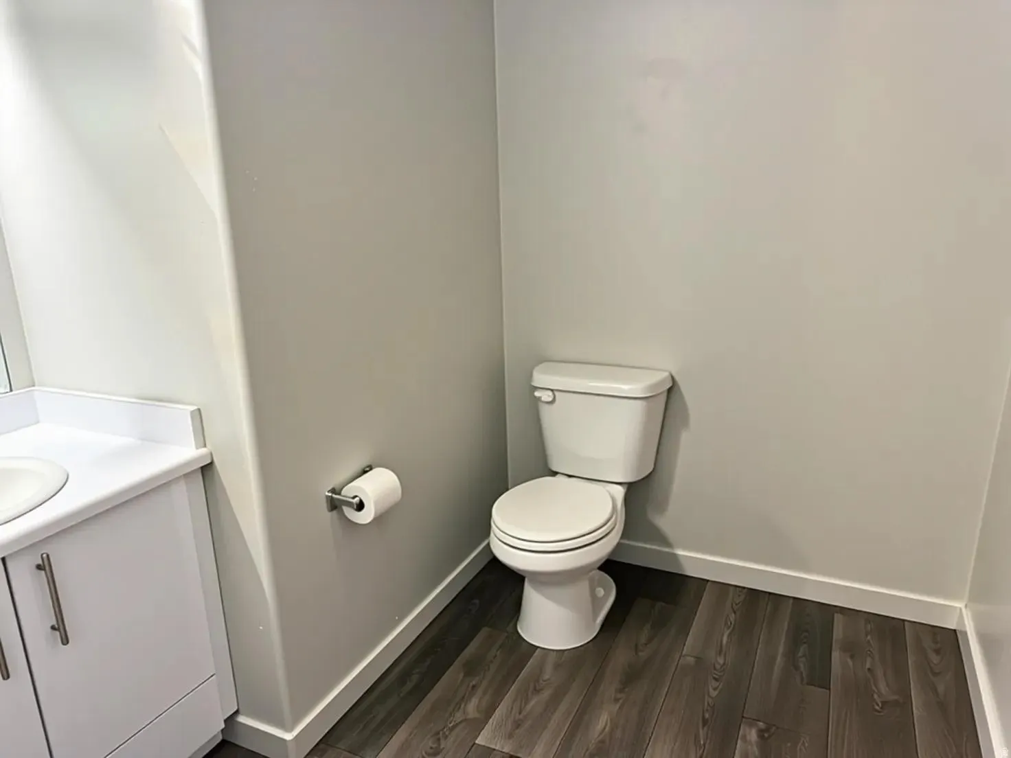 Compact bathroom featuring a white toilet, contemporary white vanity with integrated sink, brushed nickel hardware, wood-finish flooring, and crisp white baseboards