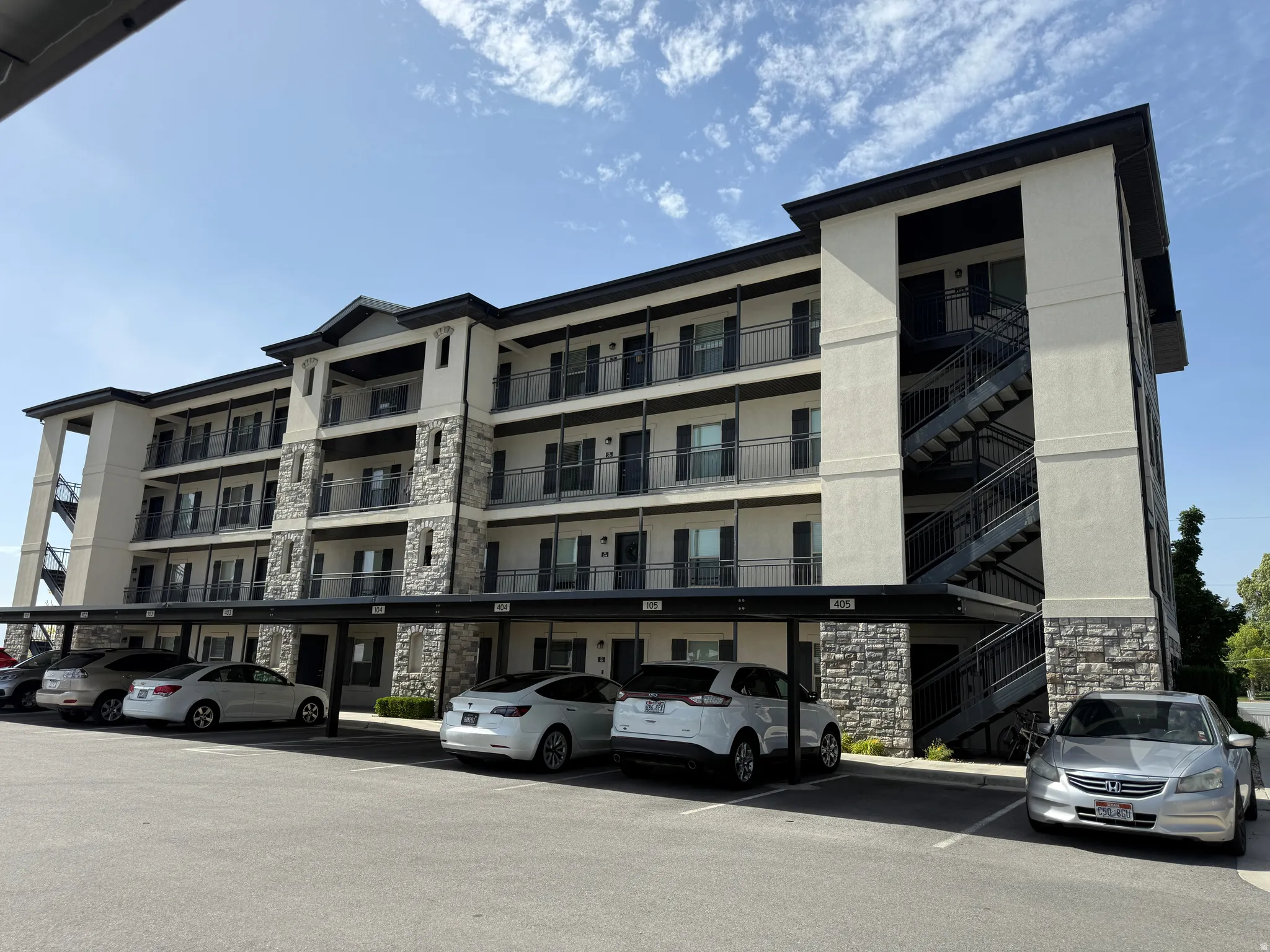 Multi-story building featuring stucco and stone veneer facade, individual balconies with black railings, exterior staircases, and covered parking