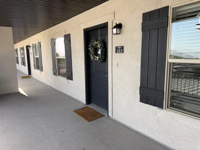 Covered exterior walkway featuring stucco facade, dark wood-finish shutters, and individual unit entry doors with exterior lighting