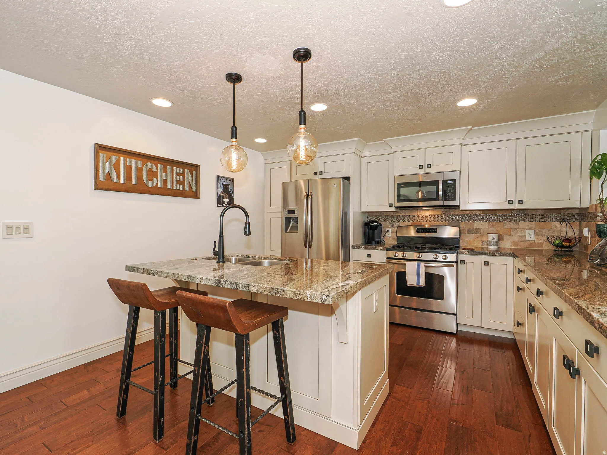 Kitchen featuring a center island with a built-in sink, pendant lighting, stainless steel appliances, white cabinetry with dark hardware, and wood-finish flooring