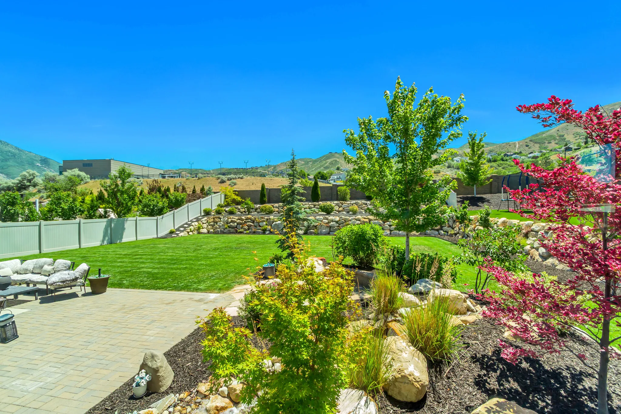 Expansive backyard featuring a paver patio, a manicured lawn, established landscaping with mature trees, and a white privacy fence