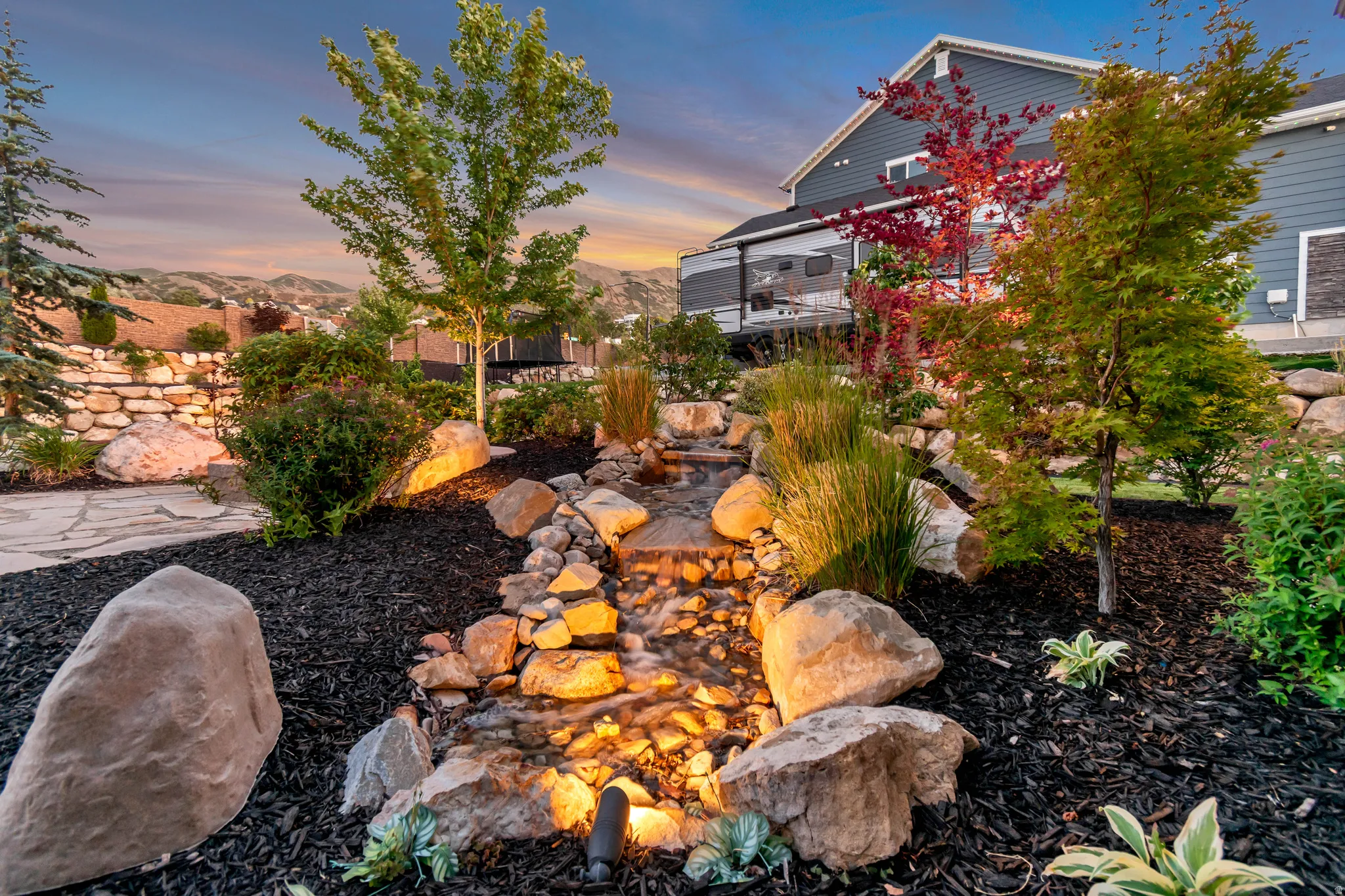 Landscaped backyard featuring a winding rock-lined water feature, mature trees, ornamental shrubs, and a dark mulch ground cover