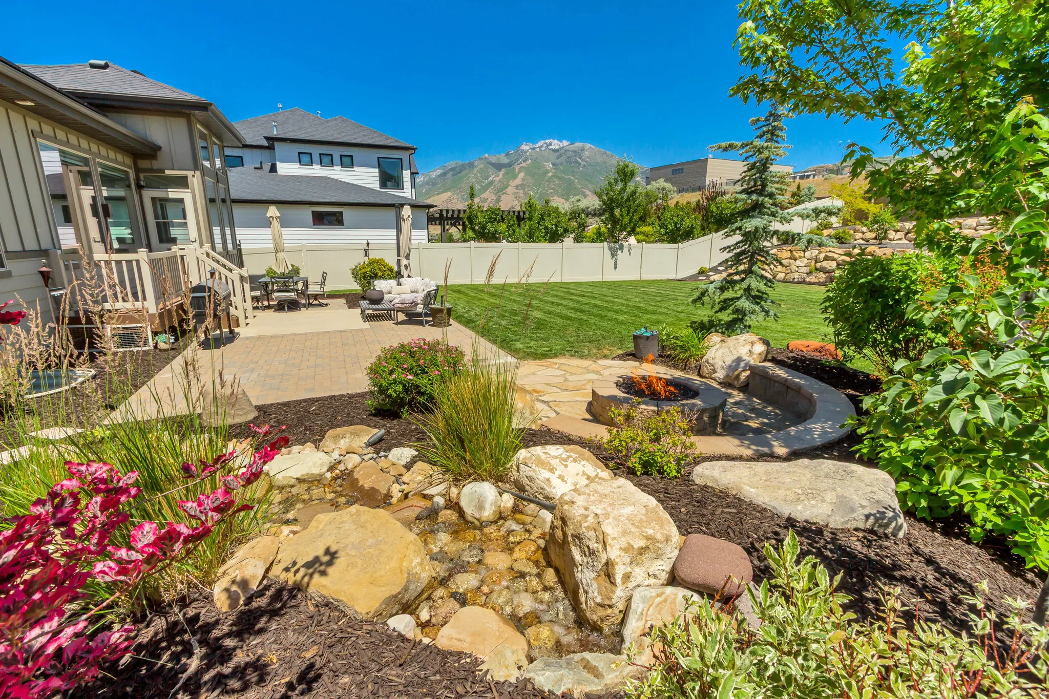 Expansive backyard featuring a stone-paved patio, manicured lawn, and a built-in stone fire pit with seating