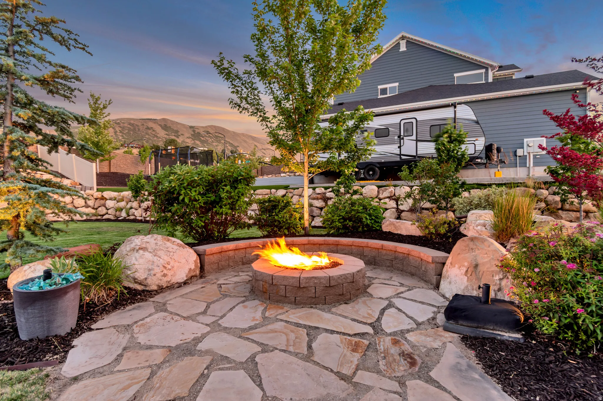 Outdoor fire pit with a natural stone patio and retaining wall, surrounded by mature landscaping and a terraced lawn