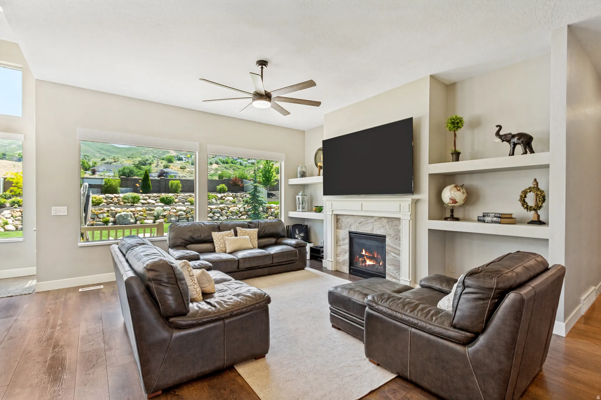 Spacious living area featuring expansive windows, a ceiling fan, wood-finish flooring, built-in shelving, and a fireplace with a stone surround and ornate mantel