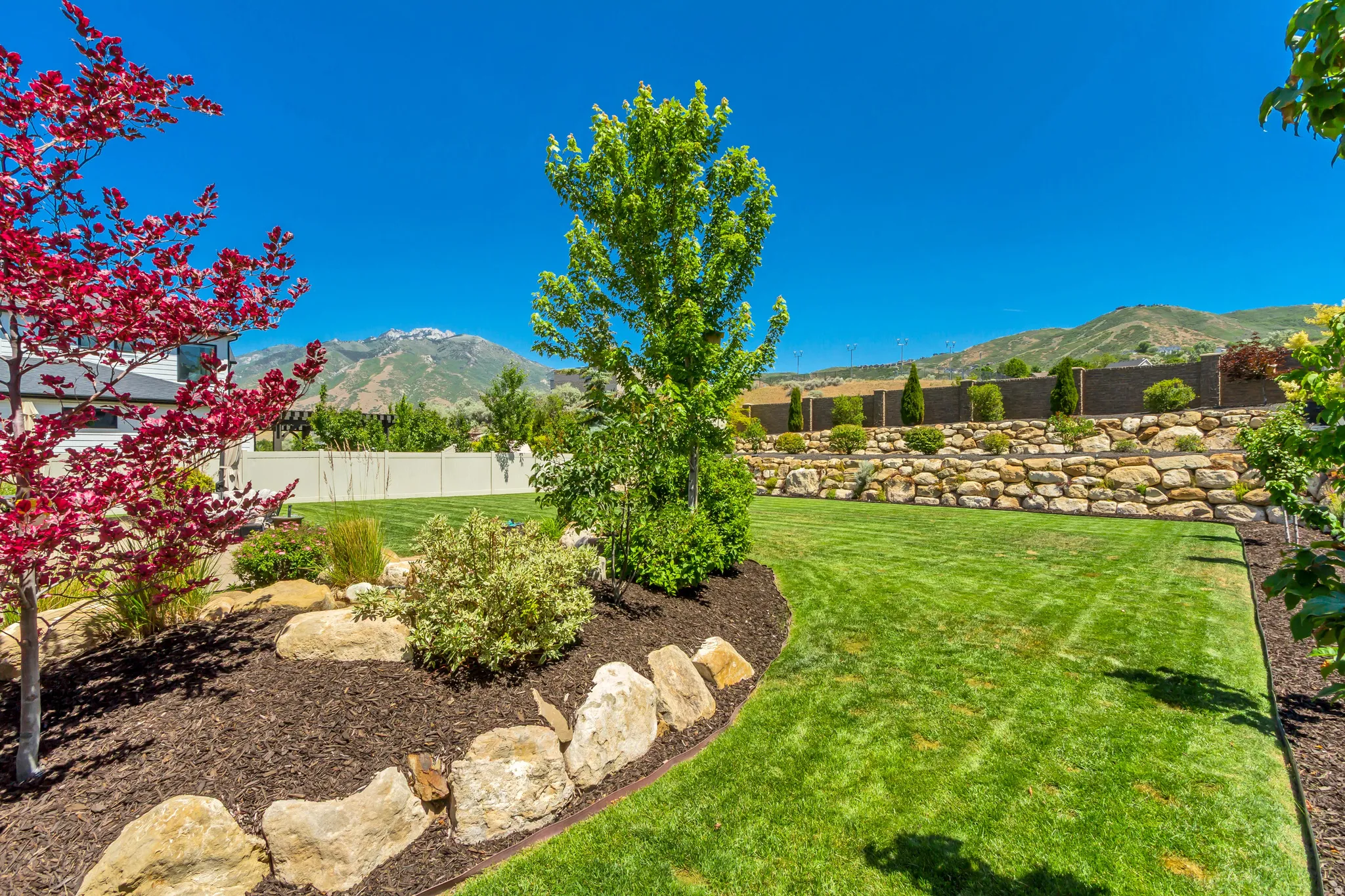Expansive backyard featuring a manicured lawn, established landscaping with mature trees and shrubs, decorative rock accents, and a tiered stone retaining wall
