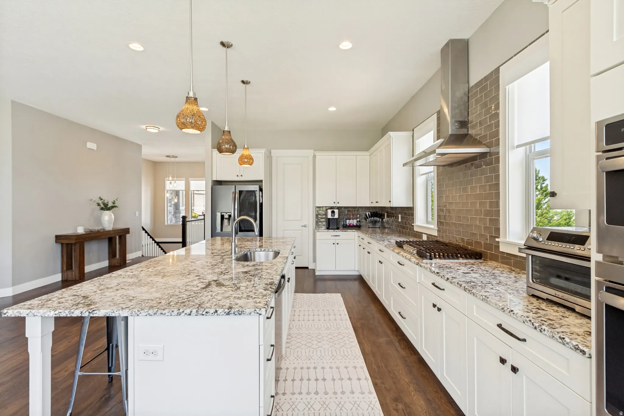 Open-concept kitchen featuring an oversized island with an undermount sink, pendant lighting, white shaker-style cabinetry, stainless steel appliances, and wood-finish flooring