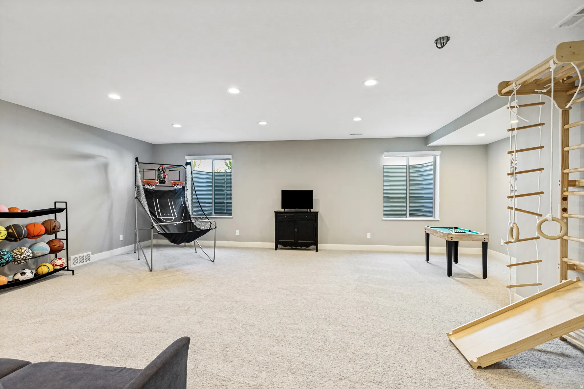 Spacious recreation room featuring light gray walls, plush carpeting, recessed lighting, and two egress windows