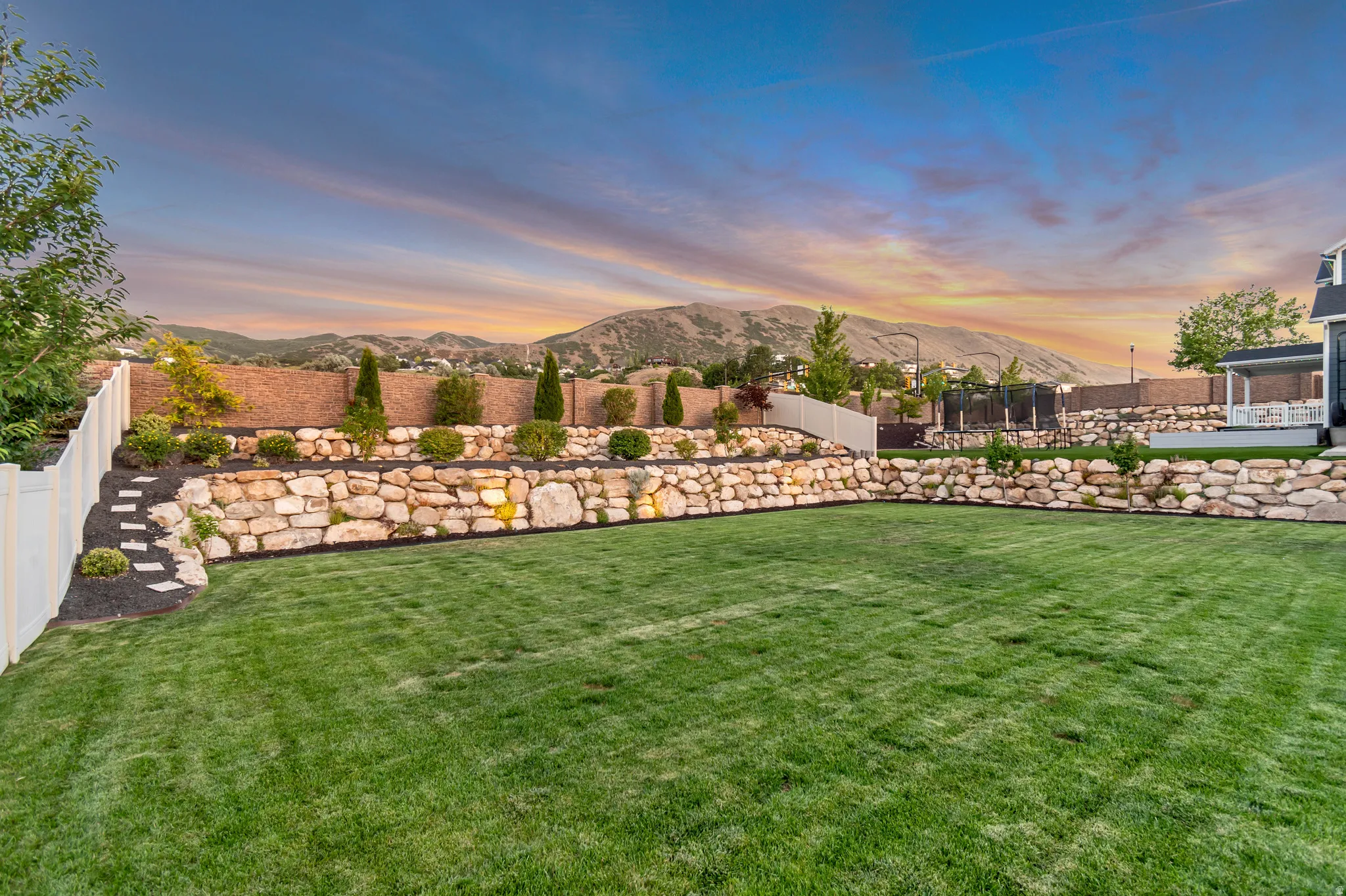Expansive lawn area featuring stacked stone retaining walls, mature landscaping, a privacy fence, and a mountain backdrop