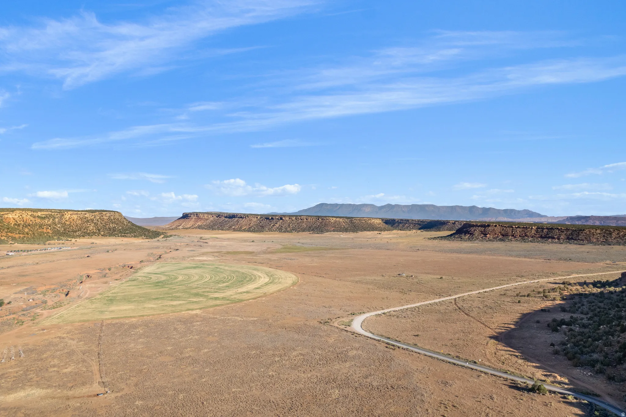 Expansive desert landscape featuring a winding dirt road, cultivated agricultural fields, and distant mesa formations under a clear blue sky