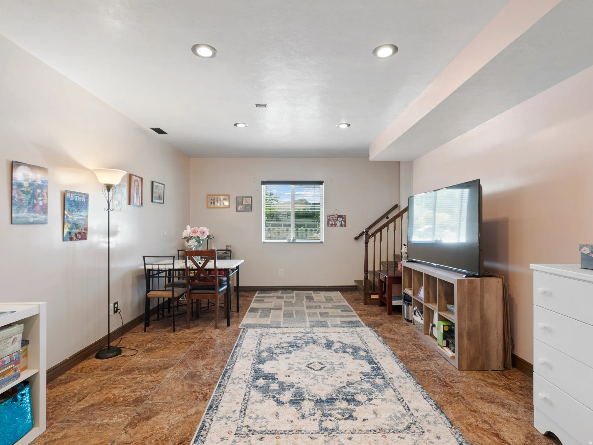 Recessed lighting illuminates an open-concept room featuring tile flooring, a window with horizontal blinds, and a wooden staircase