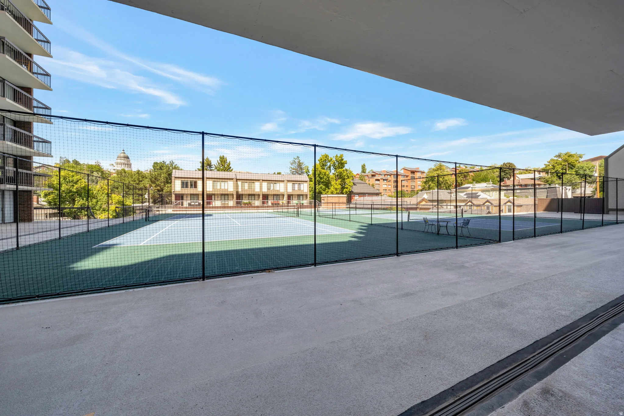 Outdoor tennis court with a light blue playing surface and green surrounds, enclosed by black chain-link fencing