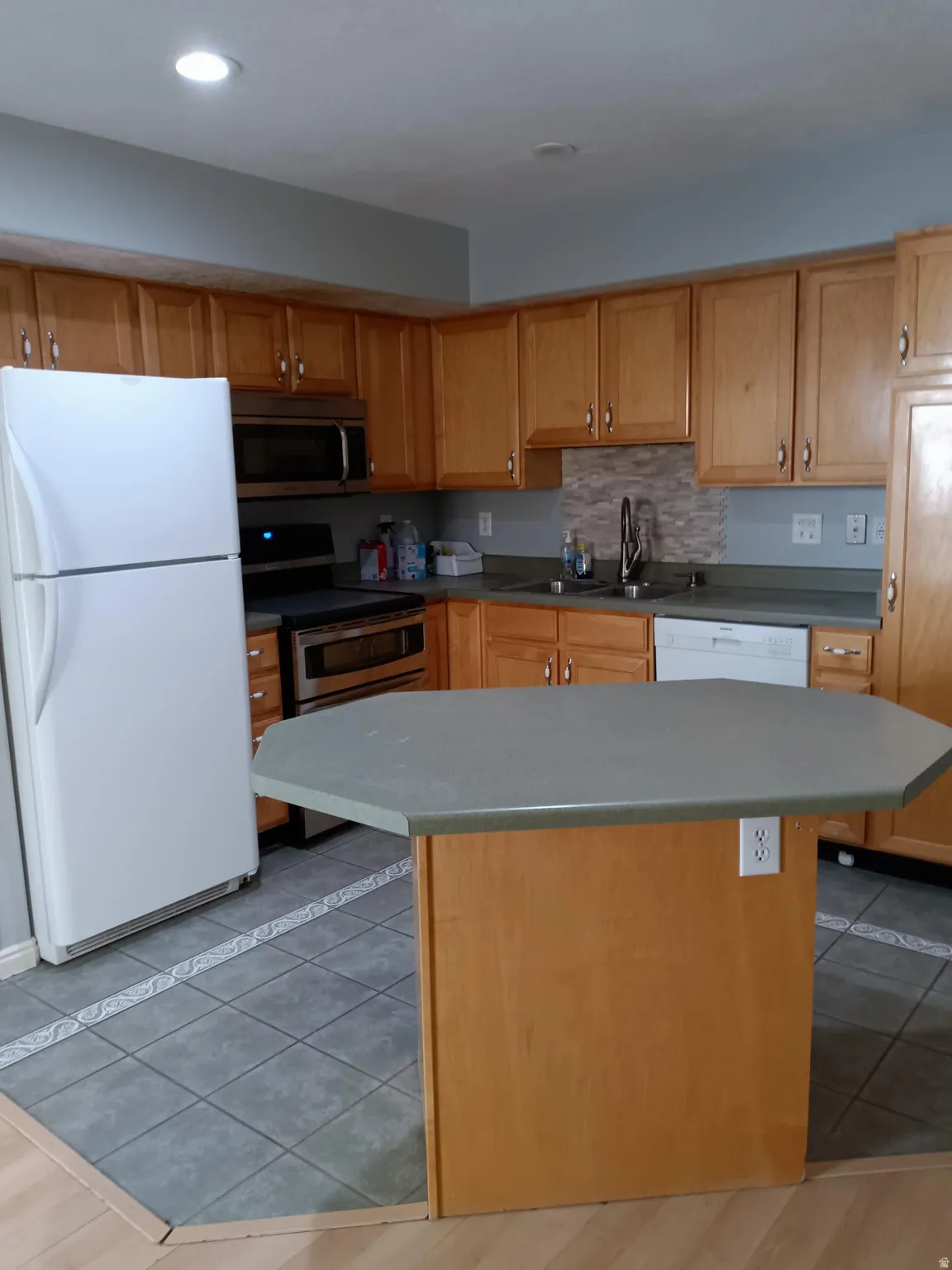 Kitchen featuring wood cabinetry, a central island with a solid surface countertop, stainless steel appliances including a built-in microwave, and a tile backsplash over the sink area