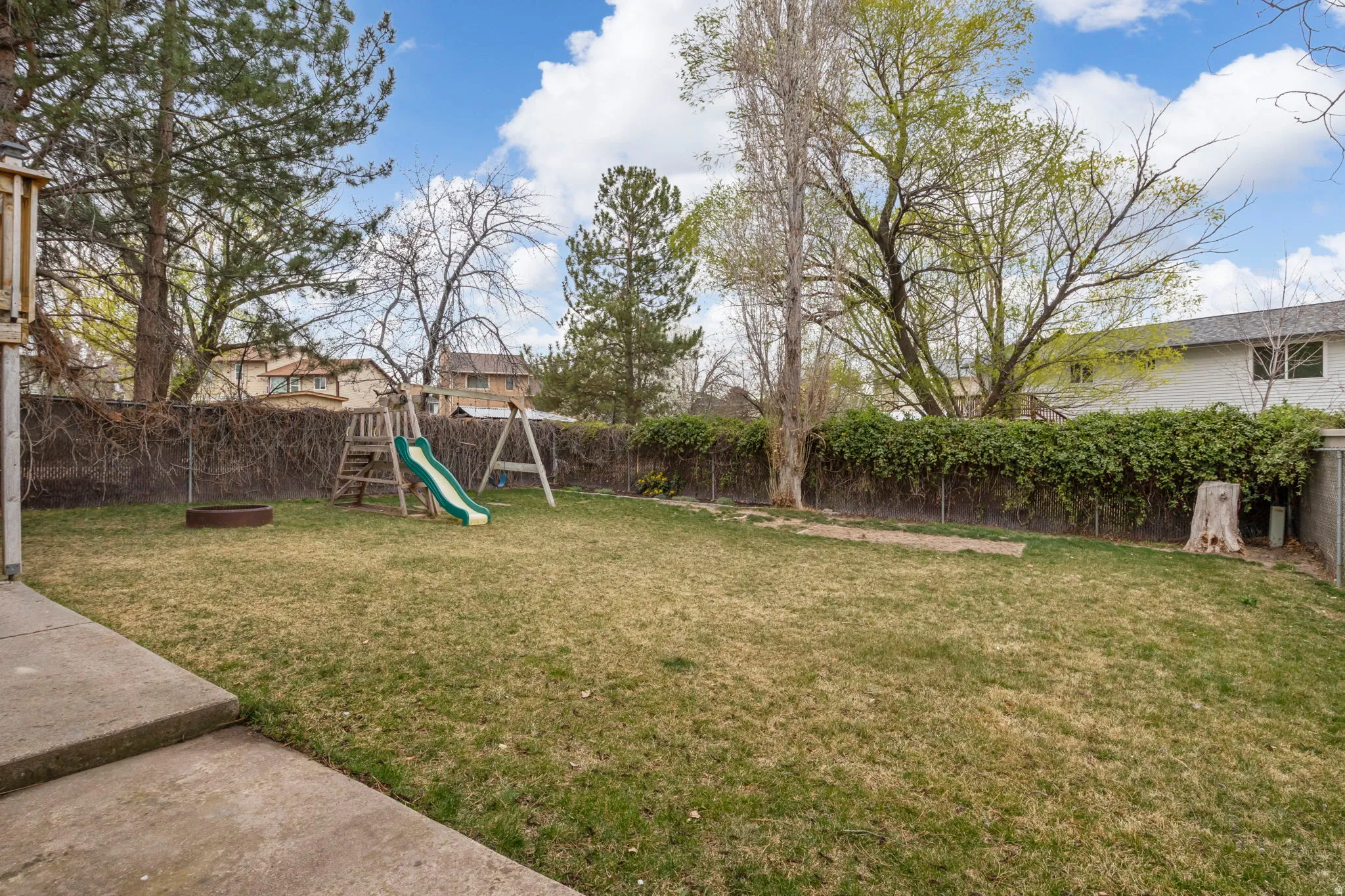 Spacious backyard featuring a sloped lawn area, mature trees, and a chain link fence with established climbing vines