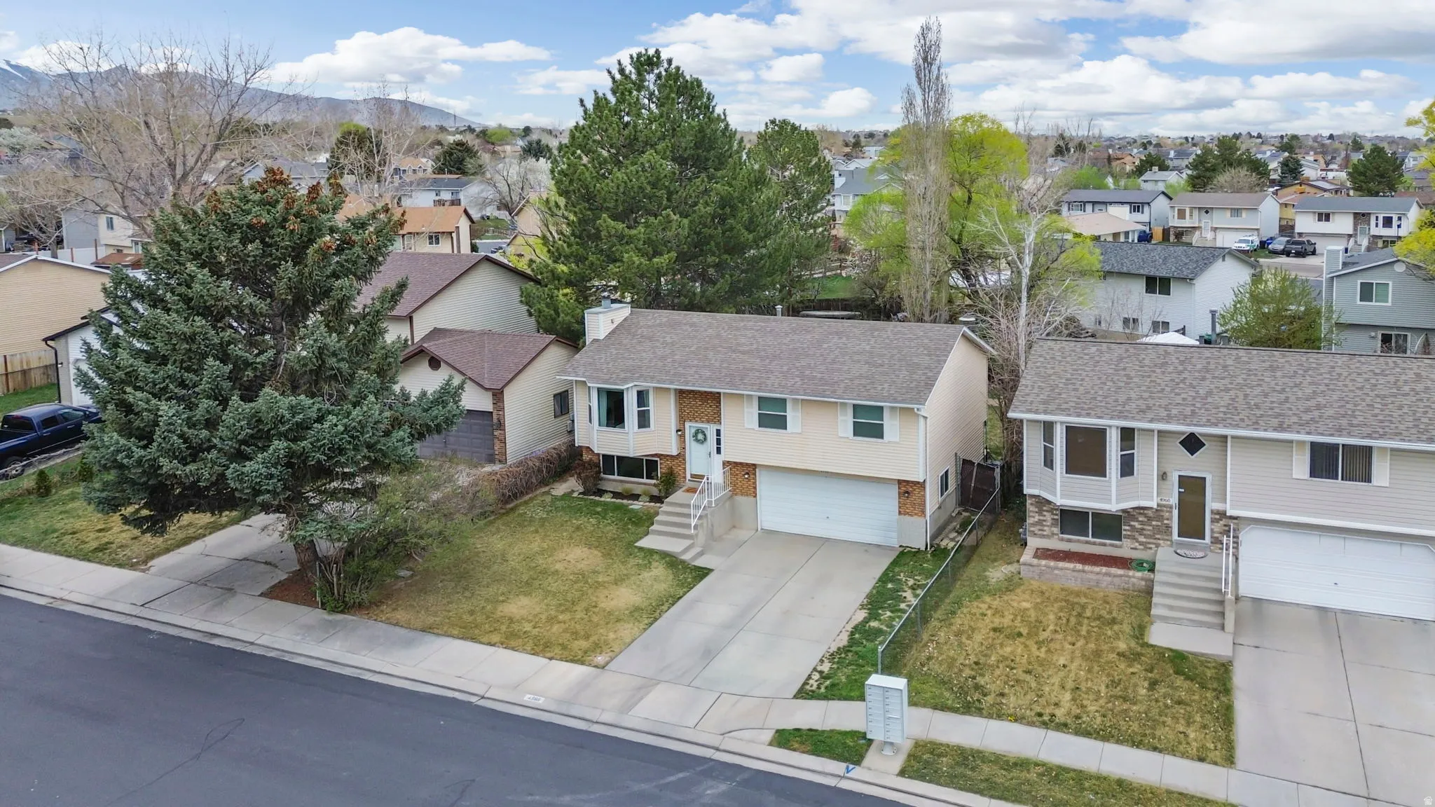 Split-level residence featuring a light-colored exterior, an attached garage, a paved driveway, and mature landscaping