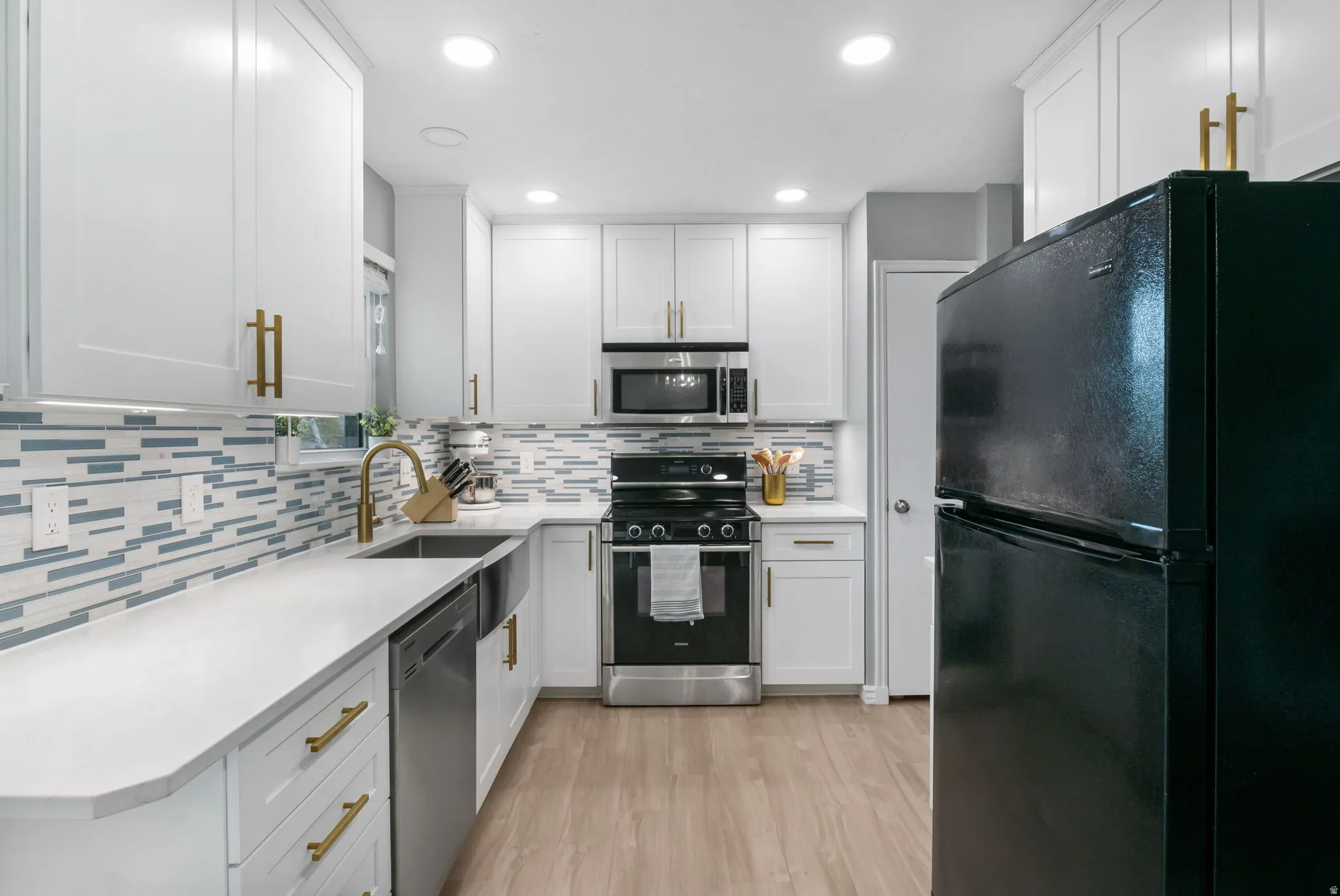 Kitchen featuring white cabinetry with gold-finish hardware, light grey countertops, a mosaic tile backsplash, stainless steel appliances, and wood-finish flooring